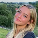 Jeune femme blonde souriante en extérieur, portant un sweat à capuche noir devant un paysage verdoyant