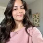 Jeune femme brune souriante portant un pull rose dans une pièce lumineuse.