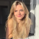 Jeune femme blonde souriante, appuyée à une fenêtre baignée par la lumière du soleil.