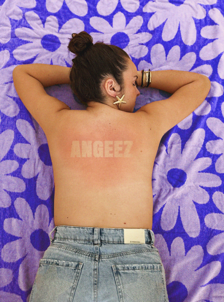 Une personne est allongée sur une serviette violette à fleurs. Son dos présente une marque de bronzage en forme du logo "ANGEEZ". Elle porte un jean bleu et une boucle d'oreille en forme d'étoile de mer.