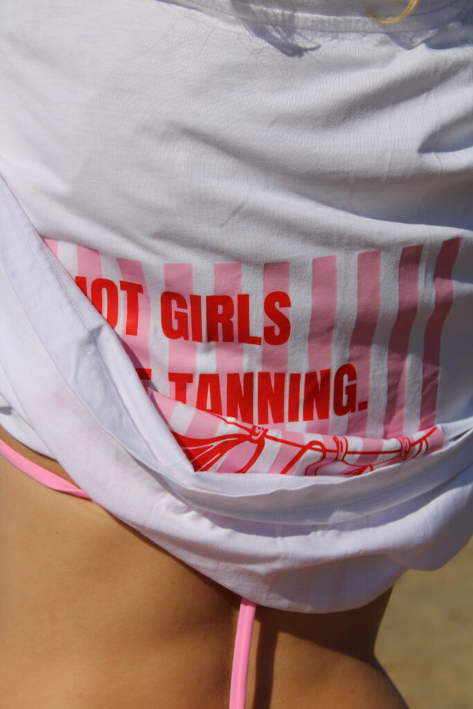 T-shirt oversize blanc avec une grande illustration rectangulaire composée de rayures roses et blanches. Une corde à linge tendue supporte deux maillots de bain rouges. Au-dessus, l'inscription « Hot Girls Like Tanning » est écrite en lettres rouges. Le logo Angeez apparaît en bas à droite du visuel.